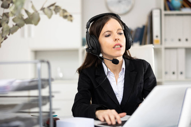 Female call center worker with headphones sitting at desk in office. - Φωτογραφία, εικόνα