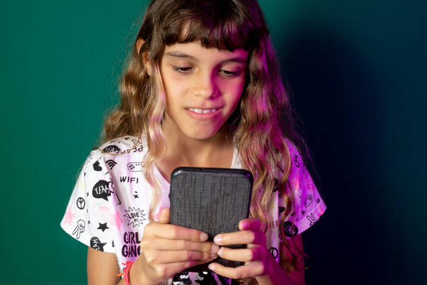 Sladká a zábavná krásná dívka se dívá na zprávy na svém mobilu. Technologie. Studio portrét se zeleným pozadím. - Fotografie, Obrázek