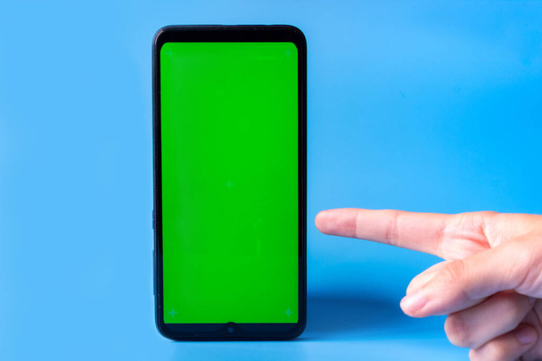 Smartphone in an upright position with a green screen on a blue background and a finger pointing at it. Chroma key. Mock up. - Foto, immagini