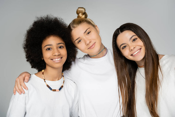 Portrait of blonde teen girl hugging smiling multiethnic friends in white t-shirts and looking at camera together while posing and standing isolated on grey, teenage friends having fun together - Photo, Image