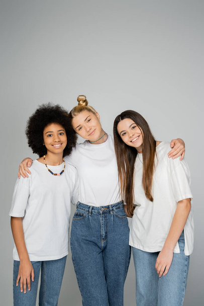Stylish and cheerful interracial teenage girls in white t-shirts and blue jeans hugging and looking at camera while standing isolated on grey, teenage friends having fun together - Photo, Image