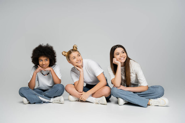 Amigas adolescentes alegres y multiétnicas en camiseta blanca y jeans azules elegantes mirando hacia otro lado mientras se sientan y pasan tiempo en un fondo gris, concepto de modelos adolescentes multiétnicos - Foto, Imagen