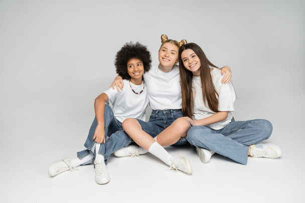 Full length of blonde teen girl hugging cheerful multiethnic girlfriends in stylish white t-shirts and jeans while looking away on grey background, multiethnic teen models concept - Photo, Image