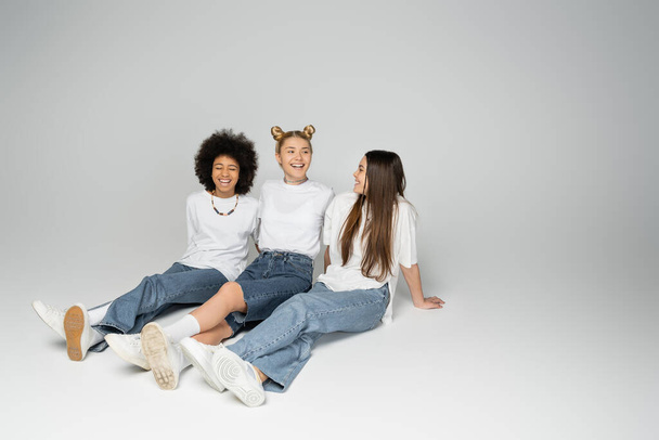 Pełna długość pozytywnych i wieloetnicznych nastolatek w białych t-shirtach i niebieskich dżinsach rozmawiających podczas siedzenia i spędzania czasu razem na szarym tle, wieloetniczne koncepcja modeli nastolatków - Zdjęcie, obraz