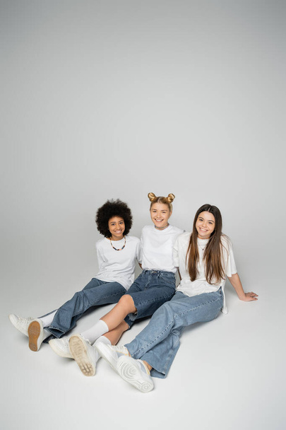 Fröhliche und multiethnische Teenager-Freundinnen in trendigen Jeans und weißen T-Shirts, die vor grauem Hintergrund sitzend in die Kamera schauen und posieren, multiethnische Teenager-Models - Foto, Bild