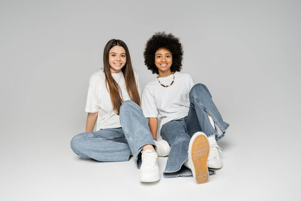 Full length of positive african american teenage girl in white t-shirt and blue jeans sitting next to brunette girlfriend on grey background, lively teenage girls concept, friendship and bonding - Foto, imagen