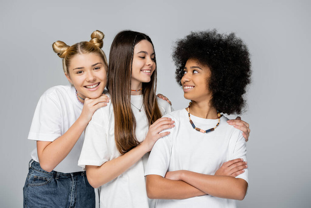 Blonde and brunette teenage girlfriends in white t-shirts hugging confident african american girlfriend crossing arms isolated on grey, lively teenage girls concept, friendship and bonding - Foto, Imagen