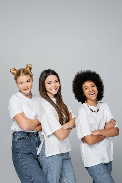 Confident and cheerful multiethnic girlfriends in casual white t-shirts and jeans crossing arms and looking at camera while standing isolated on grey, lively teenage girls concept - Photo, Image