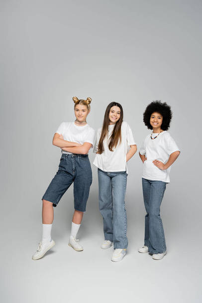Plná délka veselých a multietnických teenagerských přítelkyň v bílých tričkách a džínách pózujících společně na šedém pozadí, živé pojetí dospívajících dívek, přátelství a sbližování - Fotografie, Obrázek