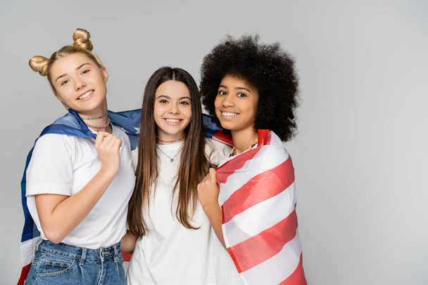 Porträt positiver und multiethnischer Teenager-Freundinnen in weißen T-Shirts, die mit amerikanischer Flagge bekleidet sind und vereinzelt in die Kamera blicken, während sie Zeit mit grauen, energischen Teenagerfreunden verbringen - Foto, Bild