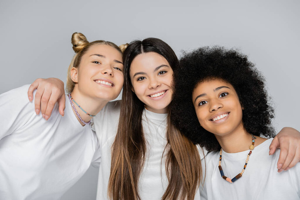 Πορτρέτο της θετικής νεαρή μελαχρινή έφηβος αγκαλιάζει διαφυλετικές φίλες σε λευκά t-shirts και κοιτάζοντας κάμερα απομονώνονται σε γκρι, ενεργητικός εφηβική μοντέλα ξοδεύουν χρόνο - Φωτογραφία, εικόνα