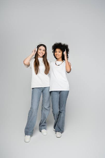 Full length of multiethnic and teenage girlfriends in white t-shirts and jeans listening music in headphones and standing on grey background, energetic teenage models spending time  - Fotografie, Obrázek