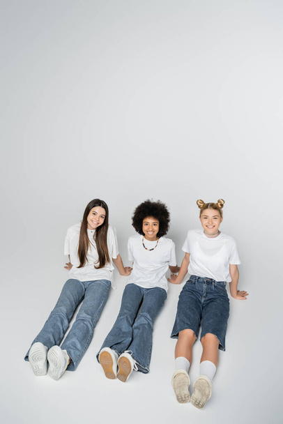 High angle view of smiling and multiethnic teenage girlfriends in white t-shirt and jeans looking at camera while sitting on grey background, adolescence models and generation z concept - Fotó, kép