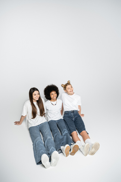 High angle view of positive interracial teenage girlfriends in white t-shirts and blue jeans looking away while sitting on grey background, adolescence models and generation z concept - Foto, Imagem