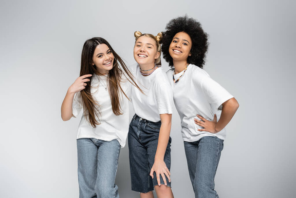 Stylish and joyful teenage girlfriends in jeans and white t-shirts looking at camera while posing together on grey background, adolescence models and generation z concept - Photo, Image