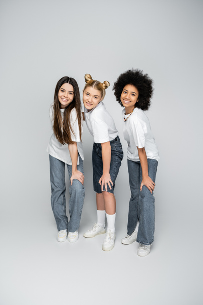Full length of stylish and teen multiethnic girlfriends in casual white t-shirts, jeans and sneakers posing together while standing on grey background, adolescence models and generation z concept - Foto, imagen