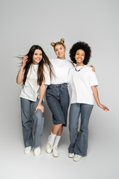 Full length of stylish teenage girl hugging multiethnic and smiling girlfriends in white t-shirts and jeans while standing on grey background, adolescence models and generation z concept - Fotoğraf, Görsel
