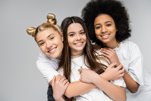 Retrato de chicas adolescentes alegres y multiétnicas en camisetas blancas abrazándose y mirando a la cámara mientras están aisladas en amigos grises y adolescentes divirtiéndose juntas - Foto, Imagen