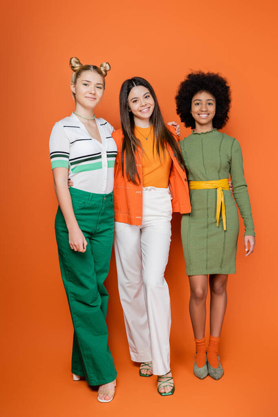 Full length of stylish and multiethnic teenage girls in trendy outfits hugging while posing and looking at camera together in orange background, teenage fashion, teen models - Foto, Imagen