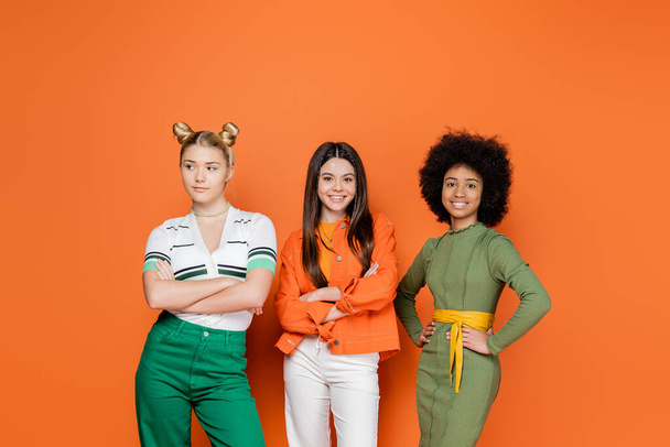 Positive and interracial teenage friends in trendy outfits crossing arms while standing and posing together on orange background, trendy generation z concept, friendship and companionship - Foto, Imagem