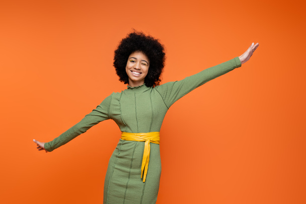 Χαρούμενη και έφηβη Αφροαμερικάνα με κομψό πράσινο φόρεμα και τολμηρό μακιγιάζ να κλείνει το μάτι και να κοιτάζει την κάμερα ενώ στέκεται απομονωμένη σε πορτοκαλί, εφηβική μόδα και τη γενιά z έννοια  - Φωτογραφία, εικόνα