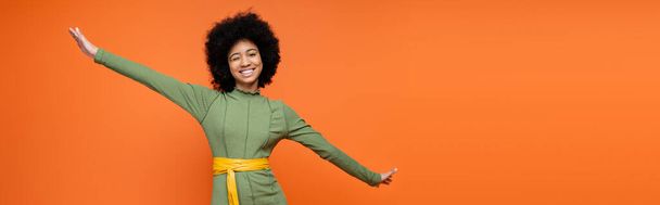 笑顔アフリカ系アメリカ人のティーンの女の子とともに大胆なメイクを身に着けています緑のドレスとウィンクでカメラ孤立でオレンジ,十代のファッションと世代zの概念,コピースペースとバナー  - 写真・画像