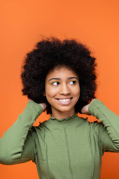 Portrait of joyful teenage african american girl with bold makeup and green dress touching hair and looking away isolated on orange, teenage fashion and generation z concept  - Fotografie, Obrázek