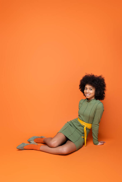Full length of positive african american teenager with bold makeup and green dress smiling at camera while sitting on orange background, youth culture and generation z concept  - Foto, imagen