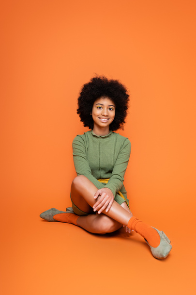 Comprimento total de alegre modelo americano africano adolescente com maquiagem colorida vestindo vestido verde e sentado em fundo laranja, cultura juvenil e conceito z geração  - Foto, Imagem