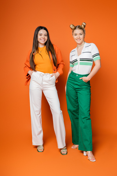 Full length of cheerful and confident brunette teenager holding hands in pockets while posing near blonde girlfriend on orange background, fashionable girls with sense of style, friendship - Zdjęcie, obraz