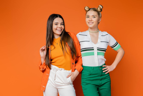 Positive brunette teenage model in casual outfit posing and standing with stylish blonde girlfriend together on orange background, fashionable girls with sense of style, friendship and bonding - Zdjęcie, obraz