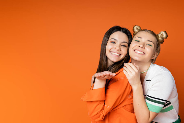 Veselá brunetka dospívající dívka s jasným make-up foukání vzduchu polibek na kameru a stojí v blízkosti módní blondýny přítelkyně izolované na oranžové, módní dívky se smyslem pro styl - Fotografie, Obrázek