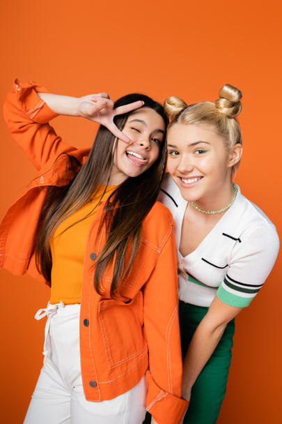 Cheerful blonde and brunette teenage girlfriends with bright makeup and stylish outfits gesturing and posing at camera while standing isolated on orange, fashionable girls with sense of style - Foto, afbeelding