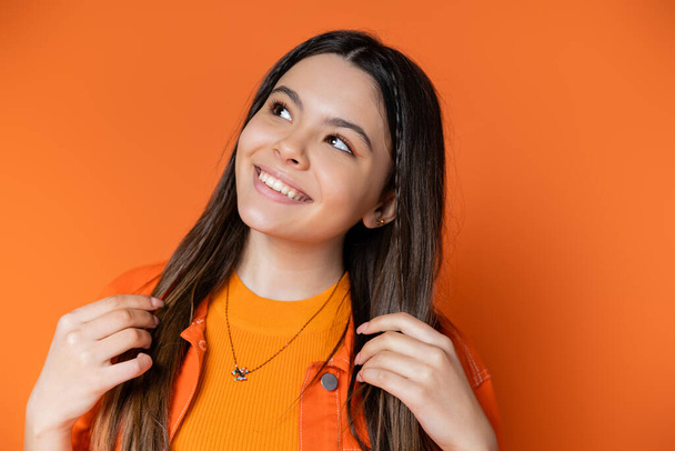 Portrait of joyful teenage model in denim jacket touching brunette hair and looking away while standing and posing isolated on orange, cool and confident teenage girl - Foto, imagen
