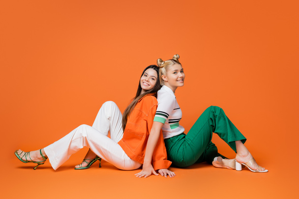 Full length of fashionable teenage girlfriends in casual outfits and heels smiling at camera while sitting back to back on orange background, cool and confident teenage girls - 写真・画像