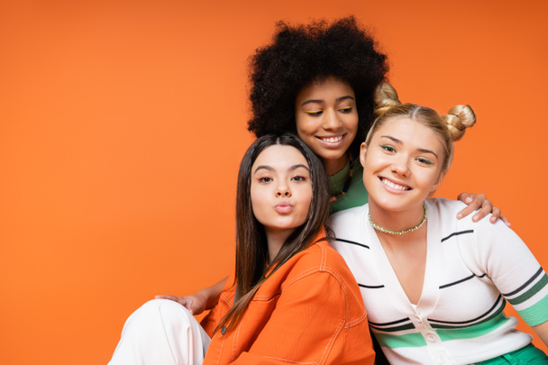Fröhliche afrikanisch-amerikanische Teenager-Mädchen mit hellem Make-up umarmt stilvolle blonde und brünette Freundinnen, während sie zusammen isoliert auf orangefarbenen, coolen und selbstbewussten Teenager-Mädchen posieren - Foto, Bild