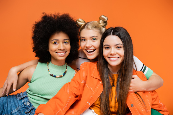 Joyful blonde teenage girl hugging fashionable and multiethnic girlfriends with bold makeup while standing isolated on orange, cool and confident multicultural teenage girls, diverse races  - Photo, Image