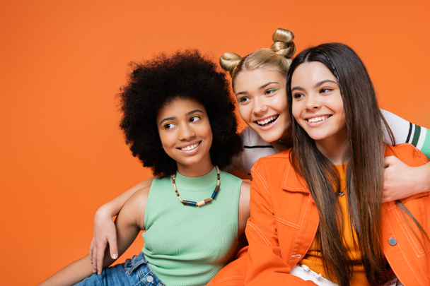 Cheerful blonde teenager with bold makeup hugging trendy interracial girlfriends and looking away together isolated on orange, cool and confident multicultural teenage girls - Foto, Imagem