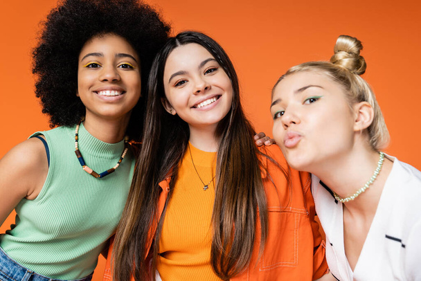 Cheerful and multiethnic teen girlfriends with bold makeup and in trendy casual clothes looking at camera while blonde friend pouting lips isolated on orange, trendy outfits and fashion-forward looks - Photo, Image
