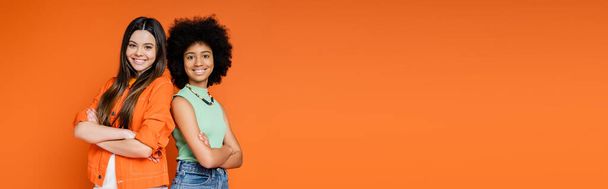 Smiling and confident multiethnic teen girlfriends with bold makeup crossing arms and standing back to back isolated on orange, teen fashionistas with impeccable style concept, banner with copy space - 写真・画像