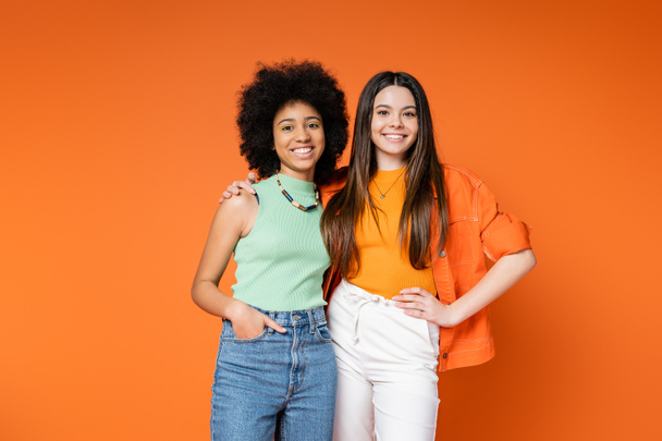 Positive and multiethnic girlfriends with bold makeup and trendy outfits hugging and looking at camera while posing isolated on orange, teen fashionistas with impeccable style concept - Φωτογραφία, εικόνα