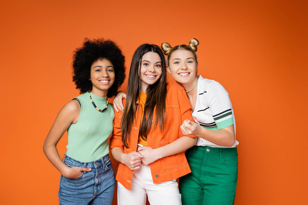 Cheerful and multiethnic teen girlfriends with bold makeup wearing trendy casual outfits hugging and posing on orange background, teen fashionistas with impeccable style concept - Photo, Image