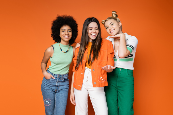 Cheerful and trendy multiethnic teenage girls with bold makeup and casual clothes posing with soap bubbles on orange background, teen fashionistas with impeccable style concept - Photo, Image