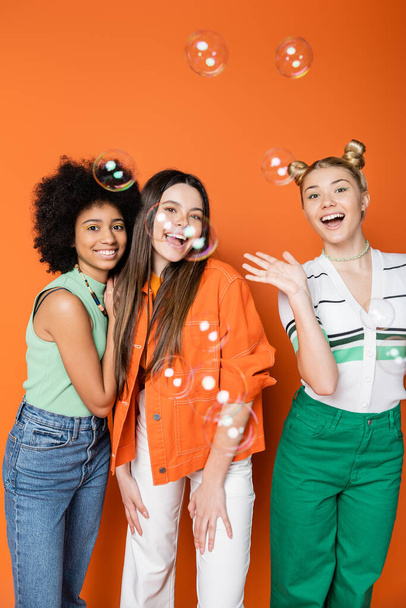 Soap bubbles near positive and interracial teenage girlfriends in casual outfits looking at camera on orange background, teen fashionistas with impeccable style concept - Foto, afbeelding