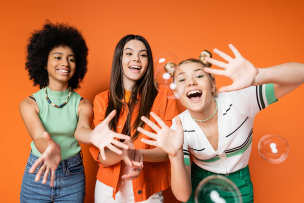 Emocionadas y alegres muchachas adolescentes multiétnicas con maquillaje audaz mirando burbujas de jabón mientras posan y de pie sobre fondo naranja, adolescentes fashionistas con un concepto de estilo impecable - Foto, imagen