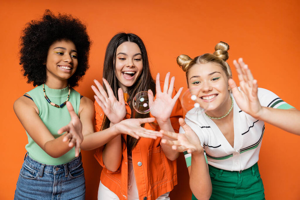 Cheerful and multiethnic teen girlfriends in stylish casual clothes looking at soap bubbles while standing on orange background, teen fashionistas with impeccable style concept - Photo, Image