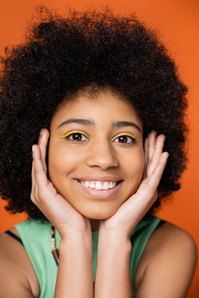 Retrato de adolescente afroamericano positivo y elegante con un maquillaje audaz tocando la cara y mirando a la cámara mientras posa aislado en naranja, chica adolescente de moda que expresa individualidad  - Foto, Imagen