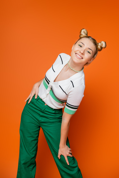 Positive und modische Teenager-Mädchen mit Frisur und kühnem Make-up posieren in lässigem Outfit und Blick in die Kamera, während sie auf orangefarbenem Hintergrund stehen, modische und trendige Kleidung - Foto, Bild