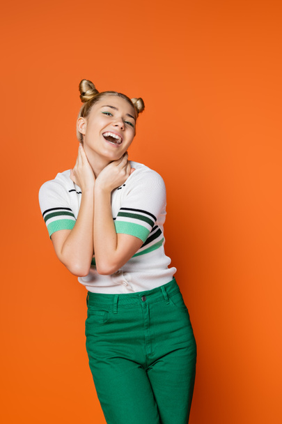 Portrait of cheerful blonde teenage girl with stylish hairstyle and bold makeup touching neck and looking at camera while posing isolated on orange, fashionable and trendy clothes - Foto, immagini