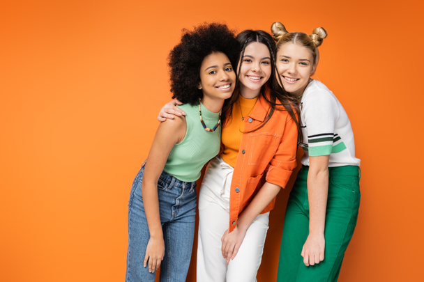 Élénk és többnemzetiségű tizenéves lányok színes smink és alkalmi ruhák ölelés és pózol együtt, miközben álló narancssárga háttér, stílusos és magabiztos pózok - Fotó, kép